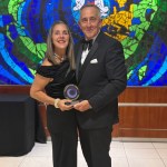 Sandra White in a black off-the-shoulder dress and her husband Martyn in a dinner suit holding her Wales Care Awards gold trophy in front of a blue and green stained glass mosaic