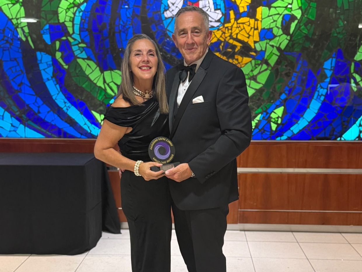 Sandra White in a black off-the-shoulder dress and her husband Martyn in a dinner suit holding her Wales Care Awards gold trophy in front of a blue and green stained glass mosaic