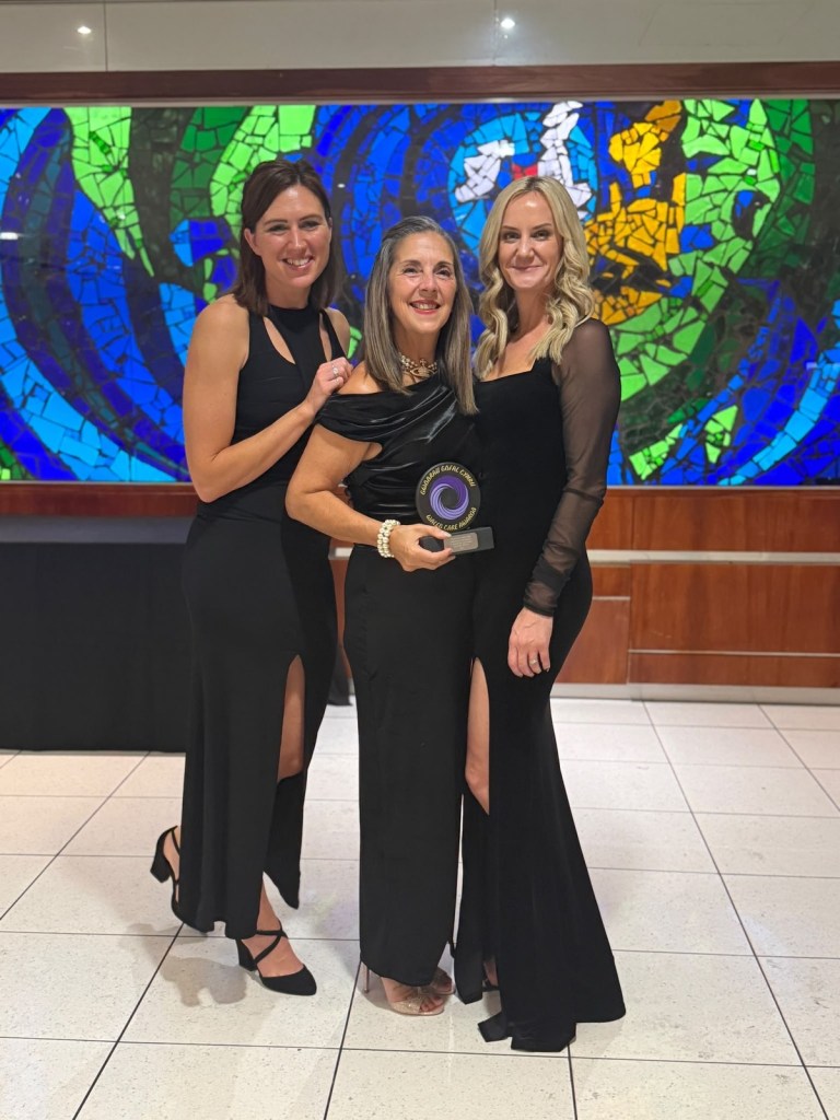 Sandra White holding her Wales Care Awards gold trophy between Megan Ludlow and Laurie Ryall of Action for Children, all in black evening dress in front of a stained glass mosaic