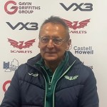 Scarlets director of rugby Nigel Davies, wearing glasses and a navy Scarlets jacket, speaks at a post-match press conference in front of a sponsor board.