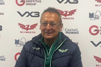 Scarlets director of rugby Nigel Davies, wearing glasses and a navy Scarlets jacket, speaks at a post-match press conference in front of a sponsor board.