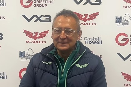 Scarlets director of rugby Nigel Davies, wearing glasses and a navy Scarlets jacket, speaks at a post-match press conference in front of a sponsor board.