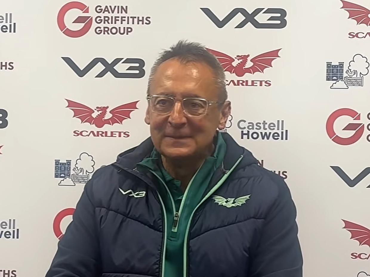 Scarlets director of rugby Nigel Davies, wearing glasses and a navy Scarlets jacket, speaks at a post-match press conference in front of a sponsor board.