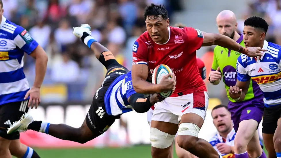 Scarlets lock Sam Lousi, in red, carries the ball through a tackle from a Stormers player in blue and white hoops.