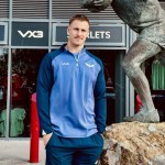 Scarlets lock Tom Allen in a blue Scarlets training top outside the club shop at Parc y Scarlets, next to a bronze statue of a rugby player