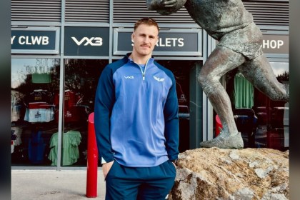 Scarlets lock Tom Allen in a blue Scarlets training top outside the club shop at Parc y Scarlets, next to a bronze statue of a rugby player