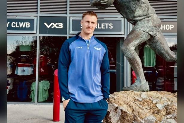 Scarlets lock Tom Allen in a blue Scarlets training top outside the club shop at Parc y Scarlets, next to a bronze statue of a rugby player