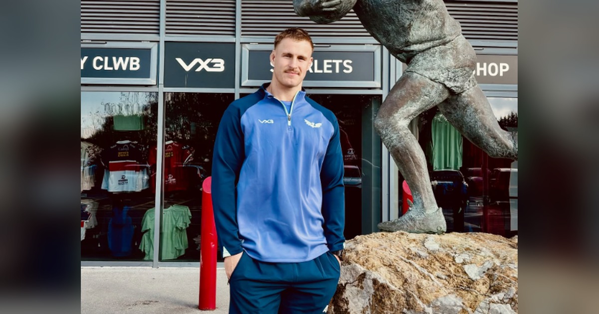 Scarlets lock Tom Allen in a blue Scarlets training top outside the club shop at Parc y Scarlets, next to a bronze statue of a rugby player