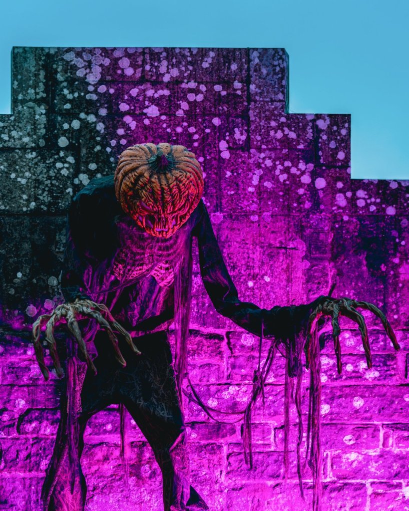 A figure with a rotting pumpkin head and long clawed hands stands against a castle wall lit in purple