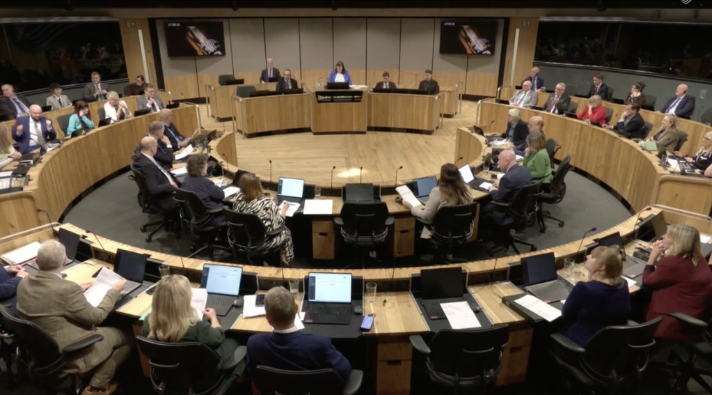 Wide view of the Senedd chamber from above, members at their desks with the Deputy Presiding Officer in the chair and the clocks on the wall screens reading 17:58