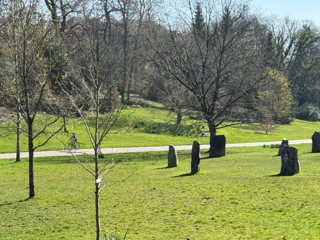 Standing stones on a grass slope in Singleton Park, Swansea, with bare trees and a cyclist on the path behind