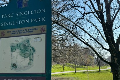 The bilingual Parc Singleton / Singleton Park sign with a City and County of Swansea crest and park map, with trees and lawns behind