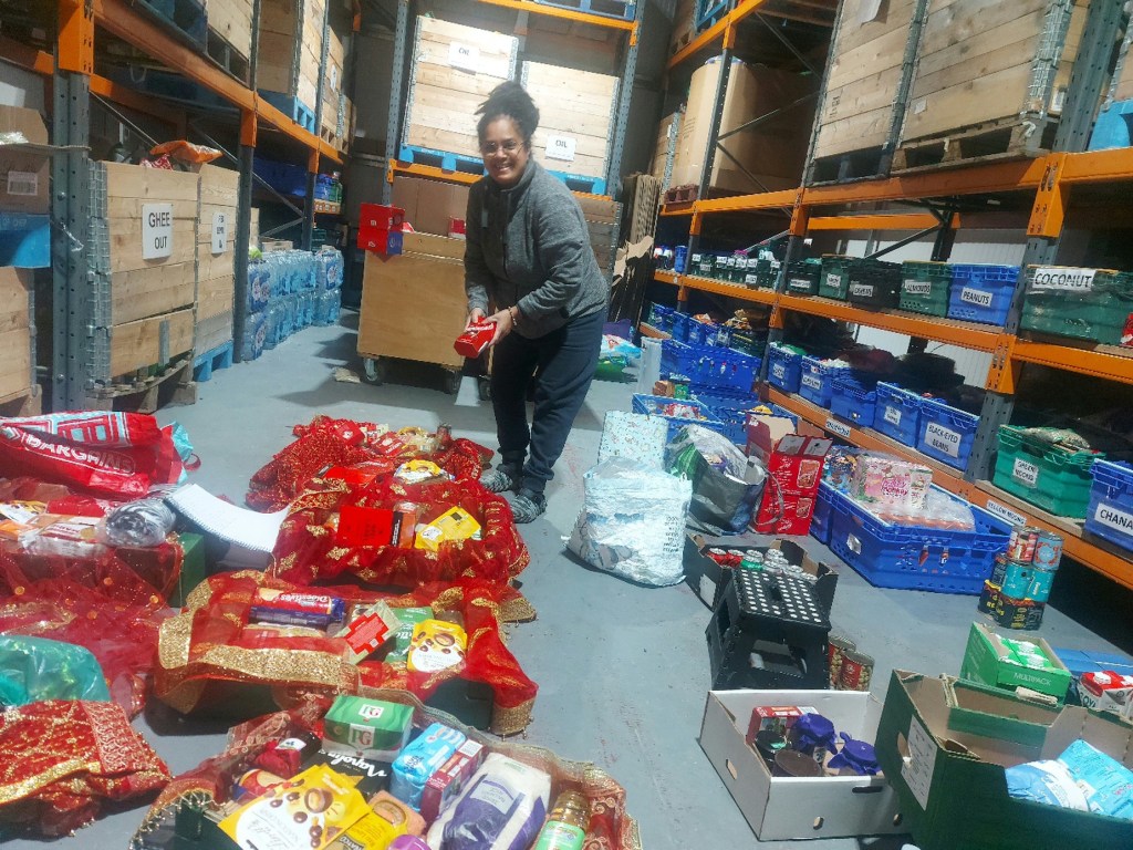 A smiling woman holds a box of food in a warehouse with labelled crates of nuts, pulses and oil on orange racking