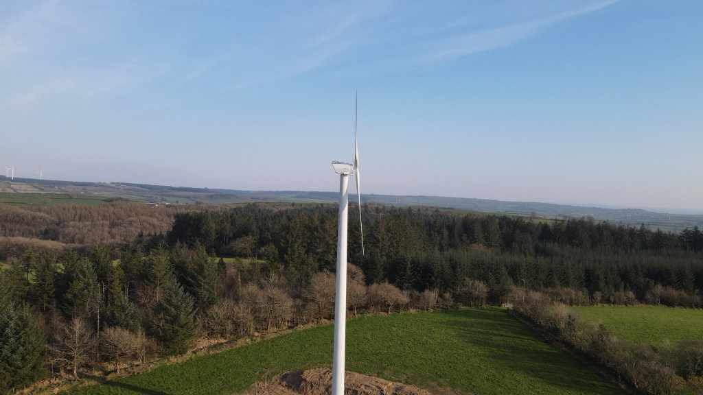 A white wind turbine standing in a field beside conifer woodland, with rolling Carmarthenshire farmland behind