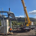 A crane lifting a curved steel section with a walkway and ladder onto a concrete pier at the Skyline base station site at Landore, with Kilvey Hill behind and a large yellow component on the ground