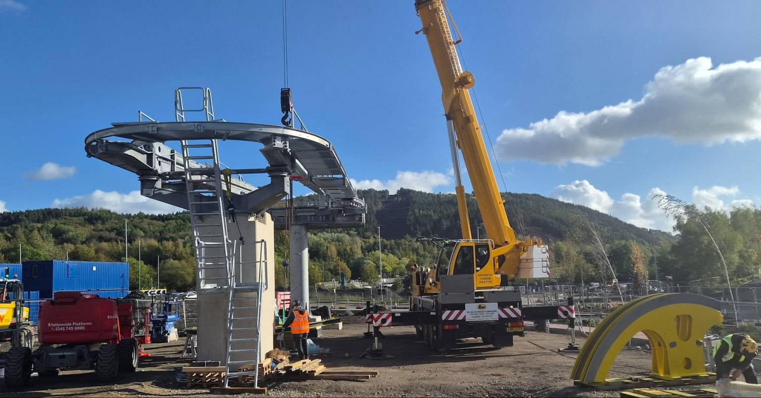 A crane lifting a curved steel section with a walkway and ladder onto a concrete pier at the Skyline base station site at Landore, with Kilvey Hill behind and a large yellow component on the ground