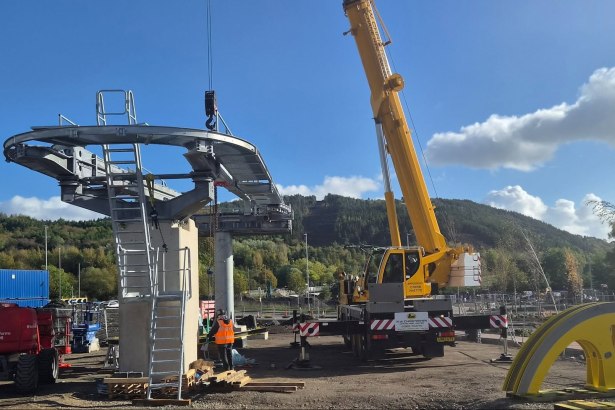 A crane lifting a curved steel section with a walkway and ladder onto a concrete pier at the Skyline base station site at Landore, with Kilvey Hill behind and a large yellow component on the ground
