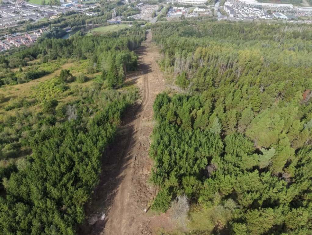 Aerial view down the cleared gondola corridor on Kilvey Hill, a strip of bare earth between conifers running towards Landore and the river