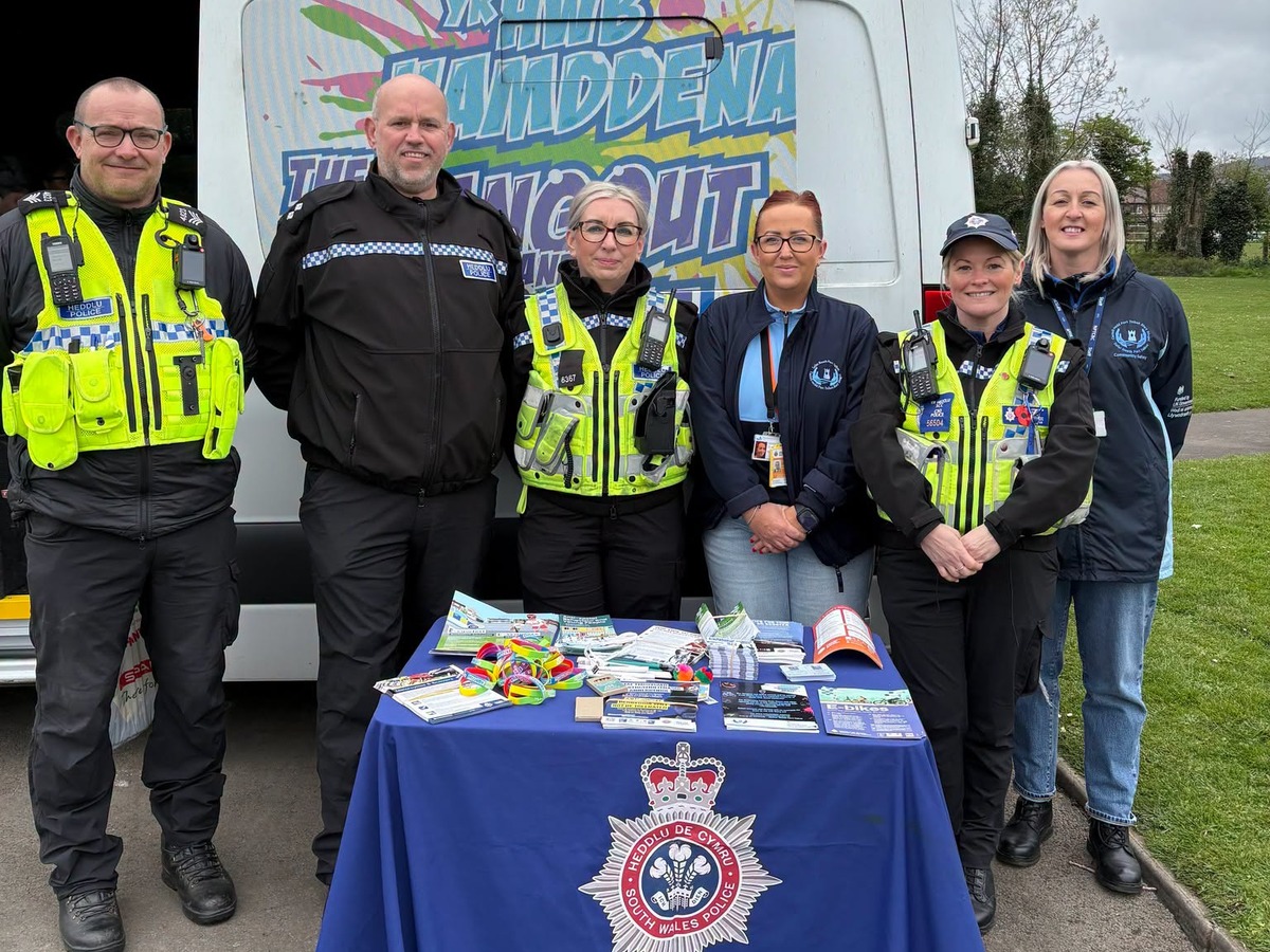 Four South Wales Police officers and two members of Neath Port Talbot Council's community safety team stand behind a police information table covered with leaflets and wristbands, in front of a youth outreach van on a path in Vivian Park.