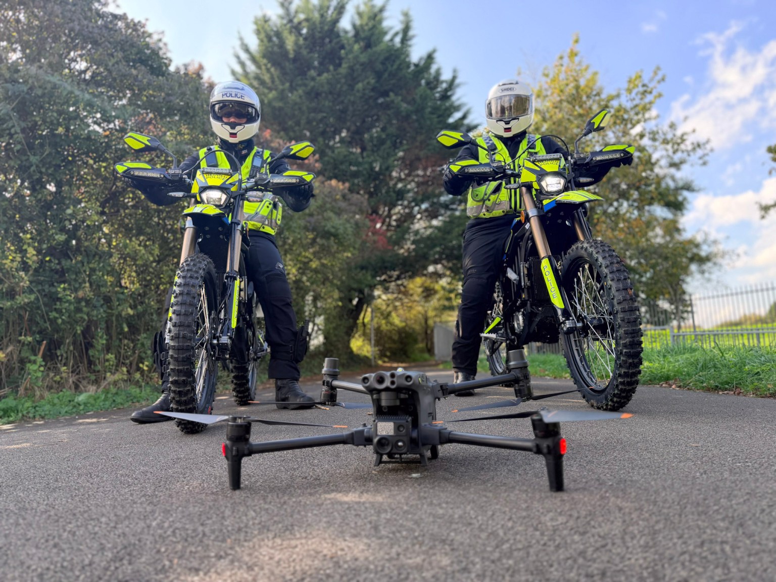 Two South Wales Police officers in helmets and hi-vis sit on yellow and black Sur-Ron electric motorbikes on a tarmac path, with a police drone on the ground in front of them