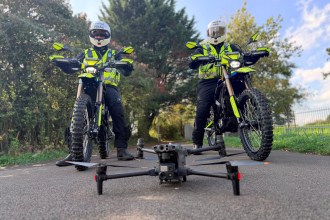 Two South Wales Police officers in helmets and hi-vis sit on yellow and black Sur-Ron electric motorbikes on a tarmac path, with a police drone on the ground in front of them