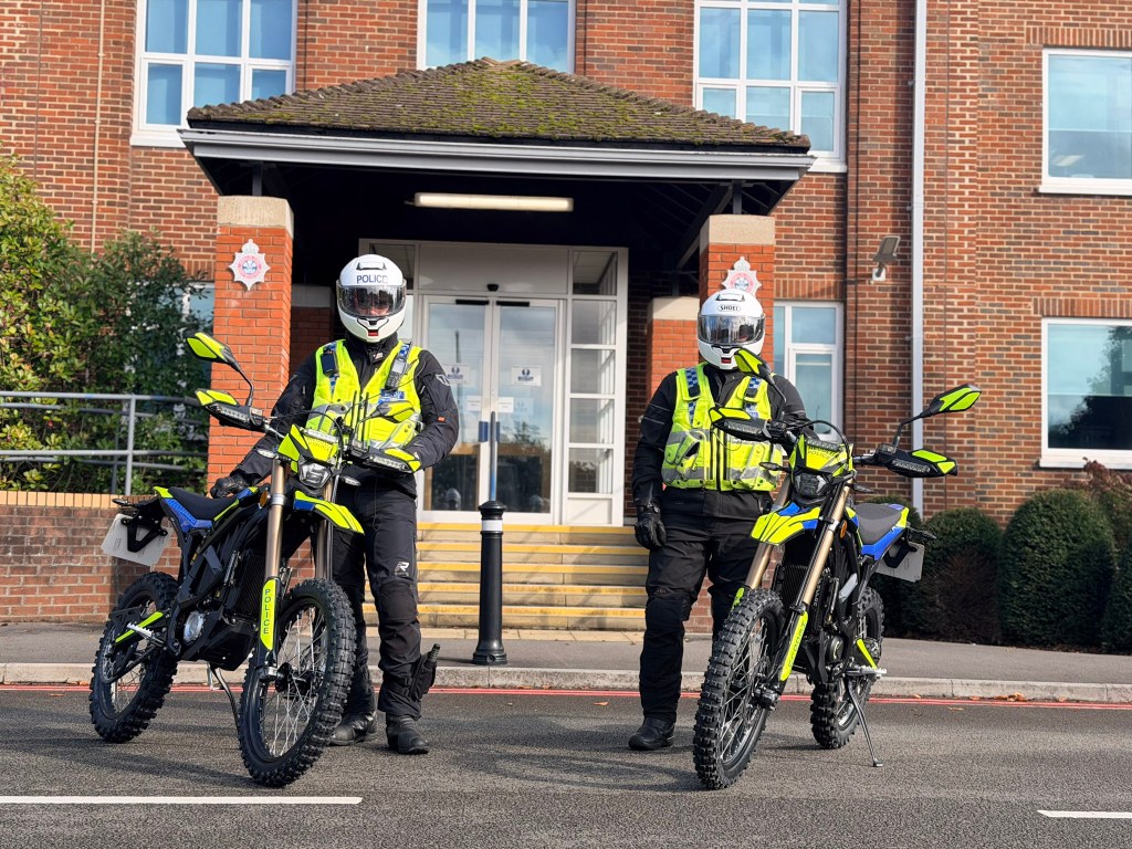 Two South Wales Police officers in helmets and hi-vis stand with yellow and black Sur-Ron electric motorbikes outside the entrance of a red-brick police building