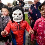 A child in a white skull mask and red jacket stands between two girls in hooded coats at a rainy Halloween event, with families behind