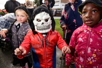 A child in a white skull mask and red jacket stands between two girls in hooded coats at a rainy Halloween event, with families behind