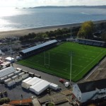 Aerial view of the rebuilt St Helen's ground with its green artificial pitch, white marquees at the Guildhall end, and Swansea Bay and the beach across Mumbles Road