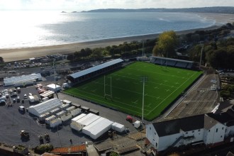 Aerial view of the rebuilt St Helen's ground with its green artificial pitch, white marquees at the Guildhall end, and Swansea Bay and the beach across Mumbles Road