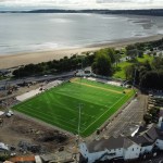 Aerial view of St Helen's in Swansea with the new green artificial pitch and floodlight columns, construction work around it, and the beach and bay towards Mumbles behind