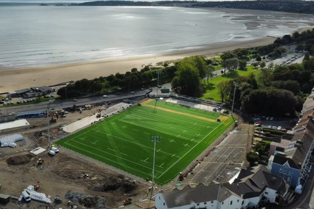 Aerial view of St Helen's in Swansea with the new green artificial pitch and floodlight columns, construction work around it, and the beach and bay towards Mumbles behind