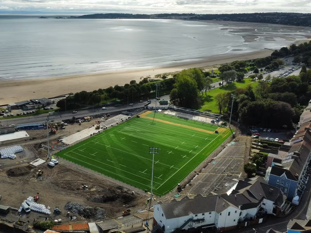 Aerial view of St Helen's in Swansea with the new green artificial pitch and floodlight columns, construction work around it, and the beach and bay towards Mumbles behind