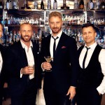 Five former Strictly Come Dancing professionals in black suits posing at a cocktail bar, one holding a martini glass