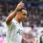 Swansea City striker Adam Idah, in a white home shirt, raises a hand to celebrate in front of the crowd at the Swansea.com Stadium