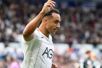 Swansea City striker Adam Idah, in a white home shirt, raises a hand to celebrate in front of the crowd at the Swansea.com Stadium