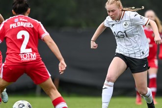 A Swansea City Women player in the white home kit runs at a Wrexham defender in red at the ThermaGlaze Ground in Garden Village