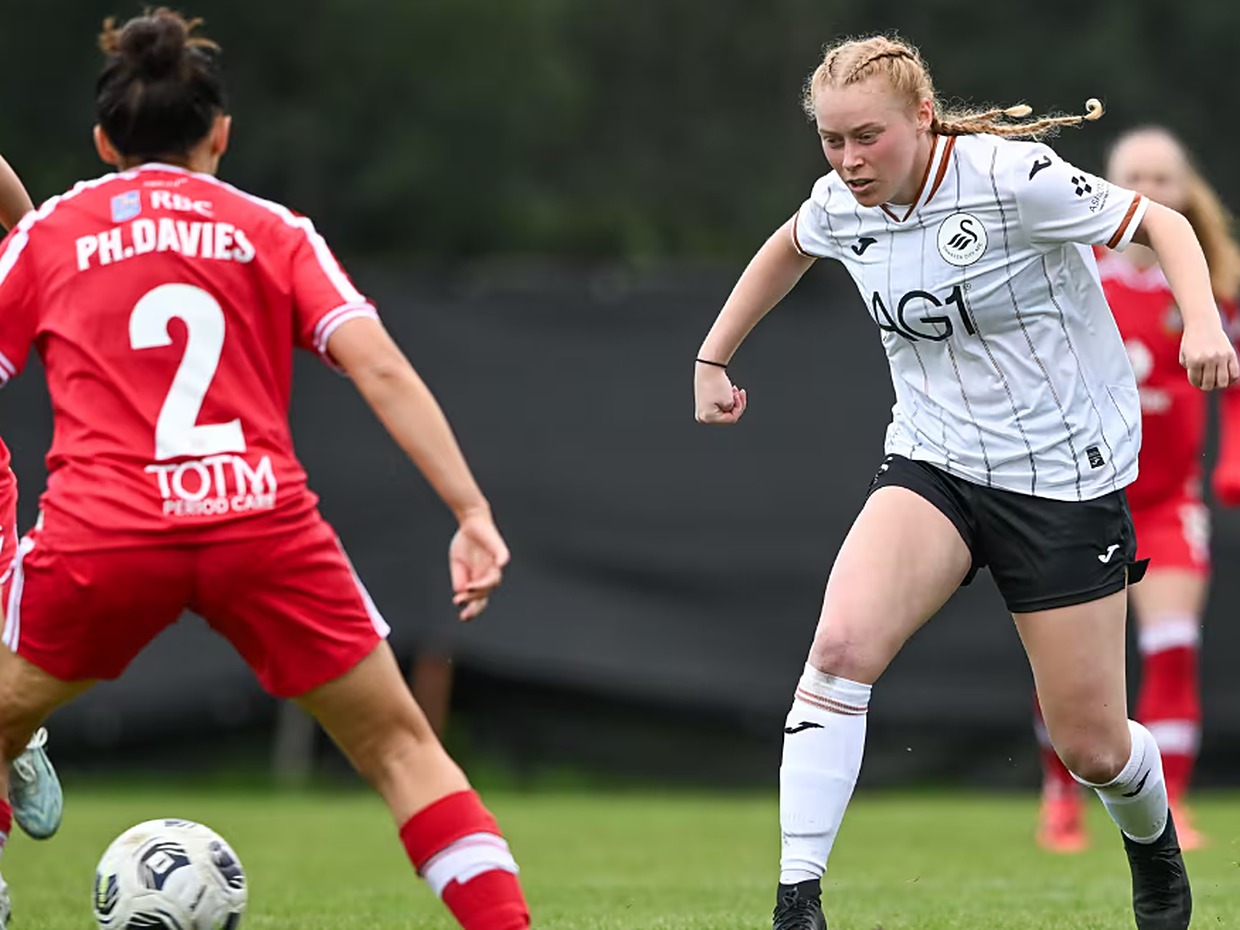 A Swansea City Women player in the white home kit runs at a Wrexham defender in red at the ThermaGlaze Ground in Garden Village