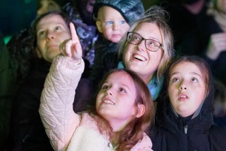 A smiling woman in glasses with three children in a crowd at night, one girl pointing up at the sky