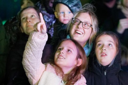 A smiling woman in glasses with three children in a crowd at night, one girl pointing up at the sky