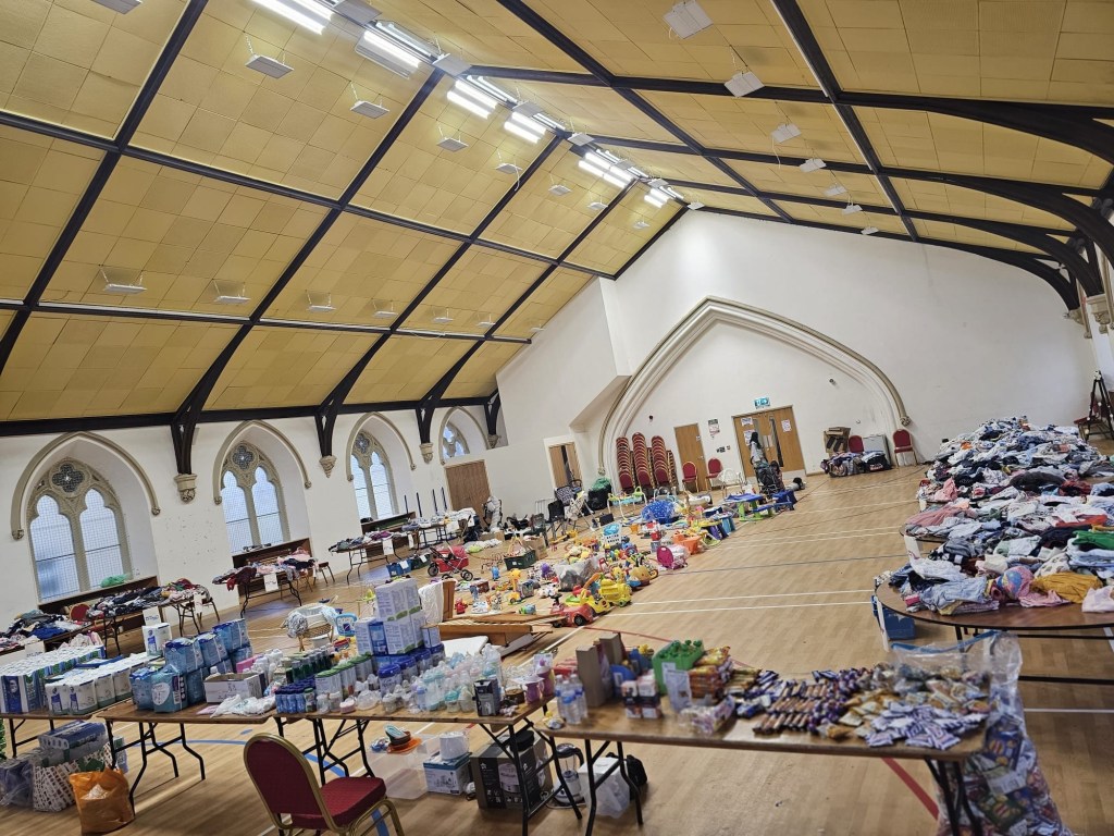 A large hall with arched windows, its floor and tables laid out with baby clothes, toys, nappies and formula