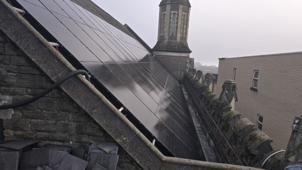 Rows of black solar panels covering a pitched slate roof beside a stone tower