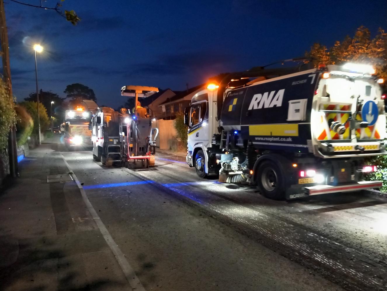 Road planer, sweeper and highways lorries working under street lights on a residential road at night during overnight resurfacing in Swansea