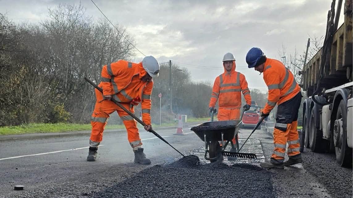 Three Swansea Council highways workers in orange hi-vis raking fresh tarmac across a pothole repair on a main road, a wheelbarrow between them