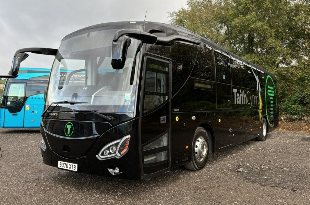 A black TaithCymru coach with green T logos parked on gravel, seen from the front