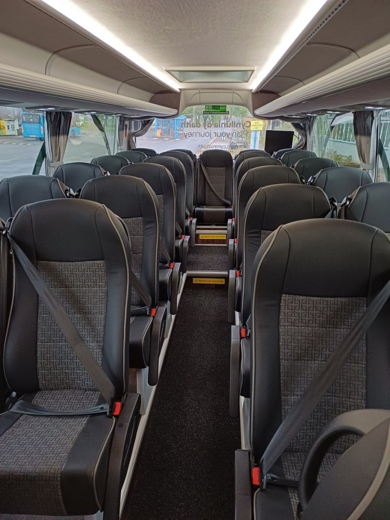The aisle of a TaithCymru coach between rows of grey and black reclining seats with seatbelts