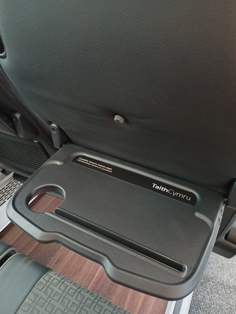 A fold-down tray table on the back of a coach seat printed with TaithCymru and Connecting Cymru in comfort