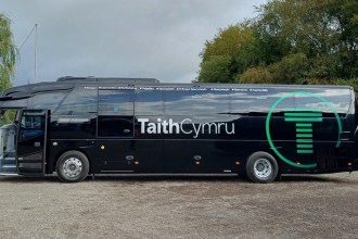 A black TaithCymru coach side-on, with its green T logo and the stops from Bangor to Caerfyrddin printed along the top