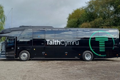 A black TaithCymru coach side-on, with its green T logo and the stops from Bangor to Caerfyrddin printed along the top