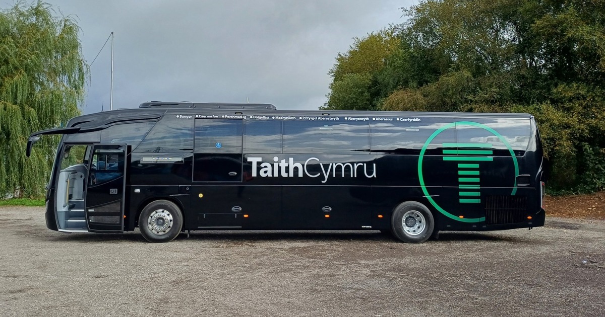 A black TaithCymru coach side-on, with its green T logo and the stops from Bangor to Caerfyrddin printed along the top